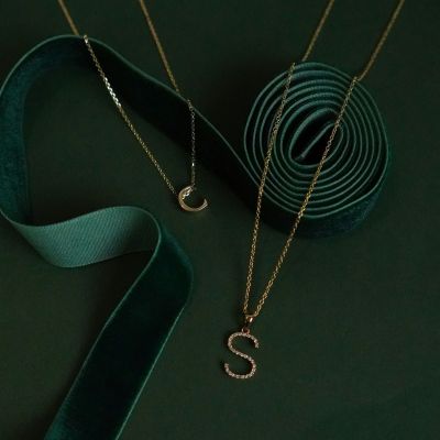 A 9ct yellow gold pendant in the shape of the letter C and a 9ct yellow gold pendant with diamonds in the shape of the lettter S are laying on a green background